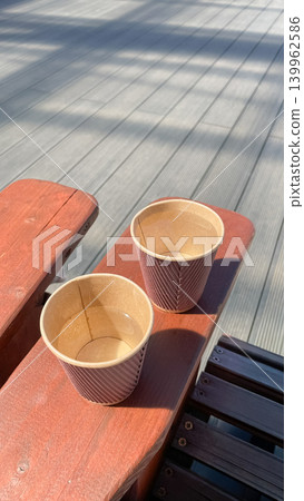 Two eco friendly paper coffee cups on a wooden bench under bright sunlight with long shadows Two eco friendly paper coffee cups on a wooden bench under bright sunlight with long shadows 139962586