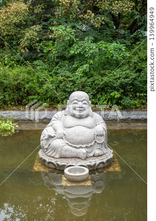 Stone Budai, or Laughing Buddha statue sitting in a shallow pond at Bongeunsa Temple in Seoul, surrounded by calm water and lush greenery. 139964949