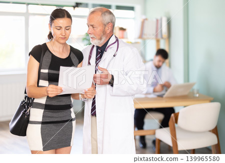 Senior male doctor and sad woman patient examine papers, discuss medical test. Senior male doctor and sad woman patient examine papers, discuss medical test. 139965580