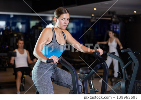 Girl performs exercises with stationary air bicycle in gym Girl performs exercises with stationary air bicycle in gym 139965693