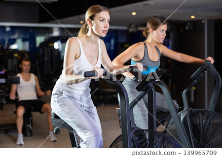 Sportive young girls doing air bike exercises in well-equipped gym Sportive young girls doing air bike exercises in well-equipped gym 139965709