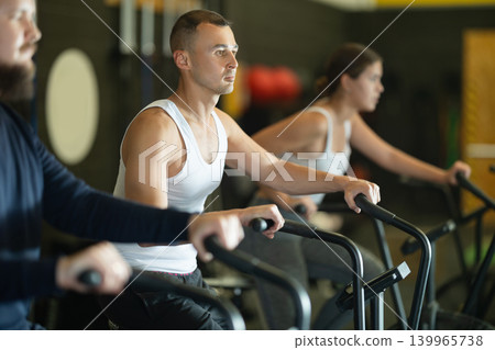 Sportive young guy doing air bike exercises in well-equipped gym Sportive young guy doing air bike exercises in well-equipped gym 139965738