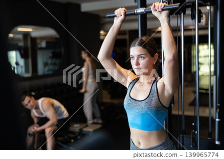 Slender athletic girl work out muscles of arms and shoulders on simulator in gym of fitness club Slender athletic girl work out muscles of arms and shoulders on simulator in gym of fitness club 139965774