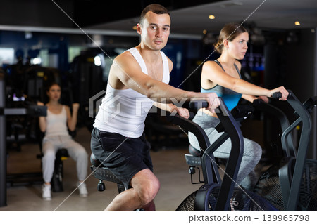 Sportive young couple doing air bike exercises in well-equipped gym Sportive young couple doing air bike exercises in well-equipped gym 139965798
