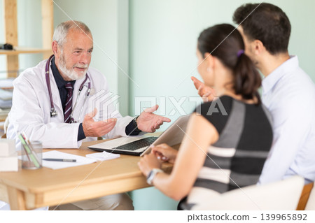 In hospital office, mature male doctor sits at table and conducts family patient consultation. In hospital office, mature male doctor sits at table and conducts family patient consultation. 139965892
