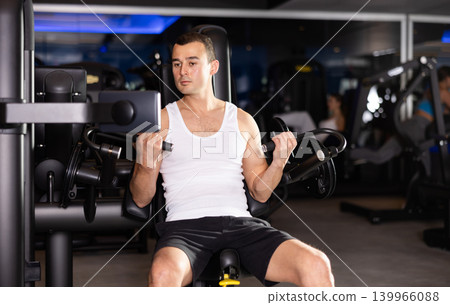 Slender athletic man work out muscles of arms and shoulders on simulator in gym of fitness club Slender athletic man work out muscles of arms and shoulders on simulator in gym of fitness club 139966088