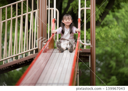一張全身照，照片中一個孩子在綠樹環繞的公園裡玩滑梯，展現了一場快樂有趣的郊遊或休閒活動。 139967043