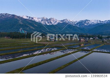 The snow-capped peaks of the Hakuba Three Peaks are reflected in the water of a rice paddy in the early morning, in Hakuba Village, Nagano Prefecture (aerial photograph taken by drone). 139969534