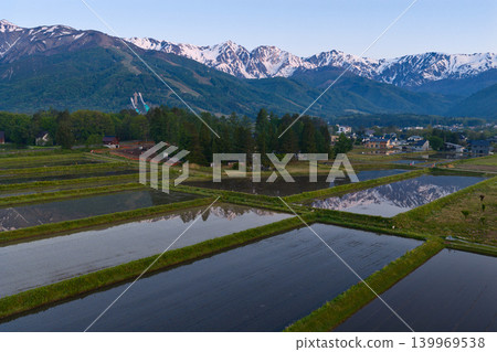The snow-capped peaks of the Hakuba Three Peaks are reflected in the water of a rice paddy in the early morning, in Hakuba Village, Nagano Prefecture (aerial photograph taken by drone). The snow-capped peaks of the Hakuba Three Peaks are reflected in the water of a rice paddy in the early morning, in Hakuba Village, Nagano Prefecture (aerial photograph taken by drone). 139969538