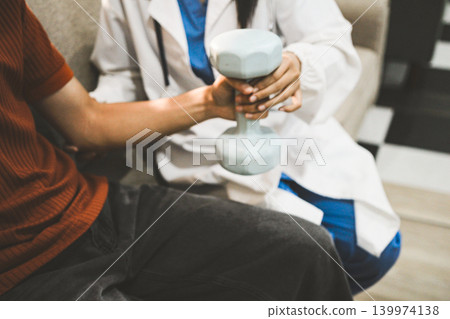 An Asian male doctor and his patient are seated in chairs, performing physical therapy using dumbbells under the doctor's supervision. The patient is experiencing pain. An Asian male doctor and his patient are seated in chairs, performing physical therapy using dumbbells under the doctor's supervision. The patient is experiencing pain. 139974138