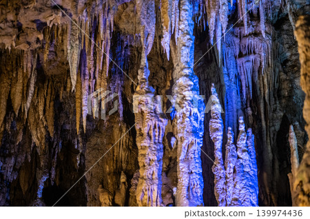 Large tall limestone rock stalagmite formations rise upward inside ancient dark underground cave interior beautifully illuminated by vibrant blue artificial scenic tourist lighting Large tall limestone rock stalagmite formations rise upward inside ancient dark underground cave interior beautifully illuminated by vibrant blue artificial scenic tourist lighting 139974436