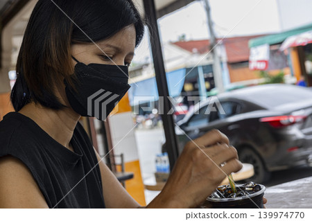 Woman Wearing Face Mask With Coffee. Asian female in black surgical mask holding coffee cup and eating at outdoor cafe, perfect for new normal lifestyle or public health themes. Woman Wearing Face Mask With Coffee. Asian female in black surgical mask holding coffee cup and eating at outdoor cafe, perfect for new normal lifestyle or public health themes. 139974770