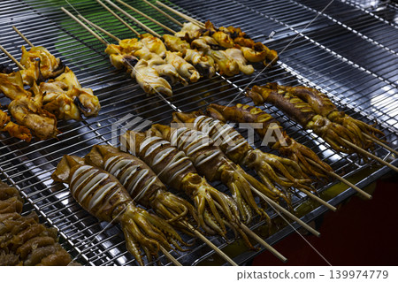 Marinated Squid Skewers On Grill Rack. Close up of seasoned seafood sticks cooking on charcoal flames with smoke, perfect for street food or asian culinary lifestyle themes. Marinated Squid Skewers On Grill Rack. Close up of seasoned seafood sticks cooking on charcoal flames with smoke, perfect for street food or asian culinary lifestyle themes. 139974779