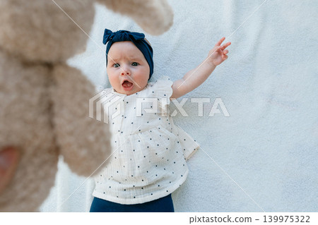 A curious and surprised baby girl looks up at a plush toy bunny being shown to her. High angle portrait of an infant child lying on a soft white blanket and reaching out with her arm A curious and surprised baby girl looks up at a plush toy bunny being shown to her. High angle portrait of an infant child lying on a soft white blanket and reaching out with her arm 139975322
