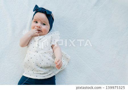 High angle shot of a sweet and calm infant girl putting her fingers in her mouth while lying on a soft white blanket. Beautiful newborn child posing in a bright, minimalist studio setting. High angle shot of a sweet and calm infant girl putting her fingers in her mouth while lying on a soft white blanket. Beautiful newborn child posing in a bright, minimalist studio setting. 139975325