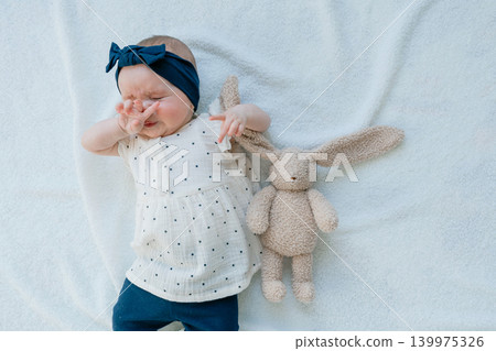 A cute and sleepy baby girl rubs her face with her hand while lying on a soft white blanket. Tired infant expressing natural emotions next to a plush toy bunny during a studio lifestyle shoot A cute and sleepy baby girl rubs her face with her hand while lying on a soft white blanket. Tired infant expressing natural emotions next to a plush toy bunny during a studio lifestyle shoot 139975326