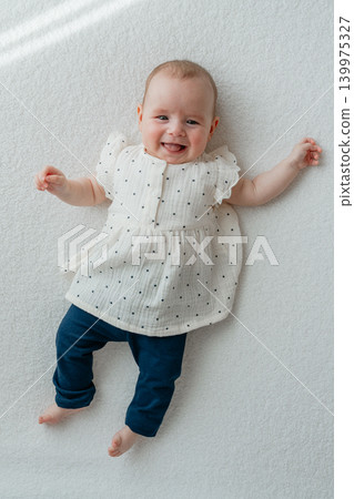 Full length portrait of a happy and smiling baby girl lying comfortably on a soft white blanket without a headband. The adorable infant is dressed in dark blue trousers and a light dotted top Full length portrait of a happy and smiling baby girl lying comfortably on a soft white blanket without a headband. The adorable infant is dressed in dark blue trousers and a light dotted top 139975327