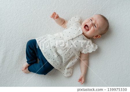A crying and upset baby girl lies on her back on a soft white blanket. Infant child expressing frustration, discomfort, colic, or hunger during a bright home or studio lifestyle photoshoot A crying and upset baby girl lies on her back on a soft white blanket. Infant child expressing frustration, discomfort, colic, or hunger during a bright home or studio lifestyle photoshoot 139975330