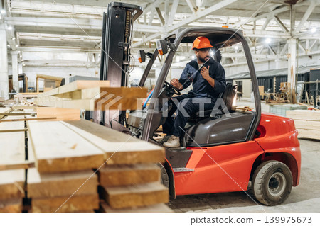 Heavy material, lifting the planks. Warehouse worker operating a forklift while handling wooden materials Heavy material, lifting the planks. Warehouse worker operating a forklift while handling wooden materials 139975673