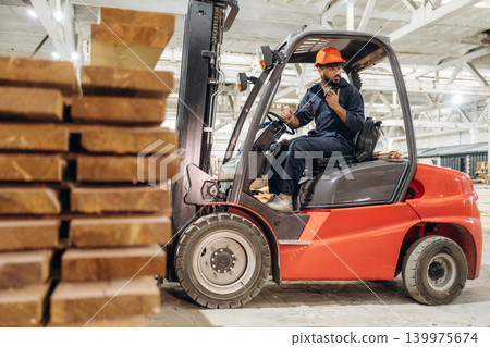 Heavy material, lifting the planks. Warehouse worker operating a forklift while handling wooden materials Heavy material, lifting the planks. Warehouse worker operating a forklift while handling wooden materials 139975674