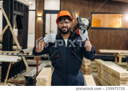 Powerful chainsaw, standing and holding it. Portrait of a male carpenter posing in a woodworking workshop 139975720