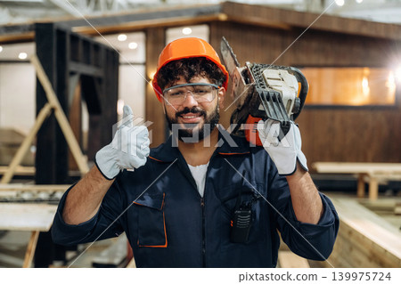 Powerful chainsaw, standing and holding it. Portrait of a male carpenter posing in a woodworking workshop Powerful chainsaw, standing and holding it. Portrait of a male carpenter posing in a woodworking workshop 139975724
