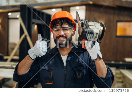 Powerful chainsaw, standing and holding it. Portrait of a male carpenter posing in a woodworking workshop Powerful chainsaw, standing and holding it. Portrait of a male carpenter posing in a woodworking workshop 139975726