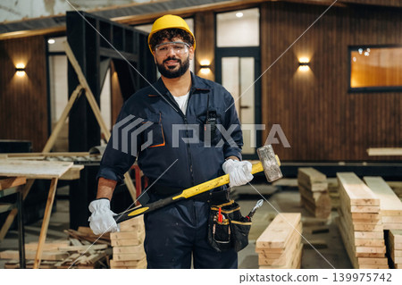Big hammer is in hands. Portrait of a male carpenter posing in a woodworking workshop Big hammer is in hands. Portrait of a male carpenter posing in a woodworking workshop 139975742