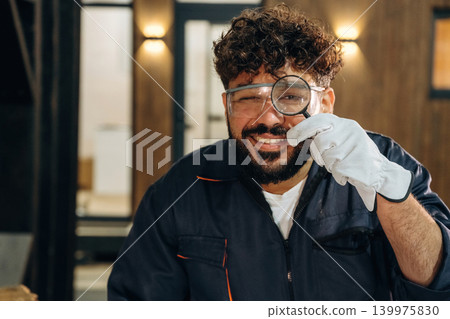 Magnifying glass, holding and looking through it. Portrait of a male carpenter posing in a woodworking workshop Magnifying glass, holding and looking through it. Portrait of a male carpenter posing in a woodworking workshop 139975830