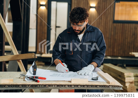 Plan is on the paper, using it. Male carpenter working with wooden materials in a professional workshop Plan is on the paper, using it. Male carpenter working with wooden materials in a professional workshop 139975831