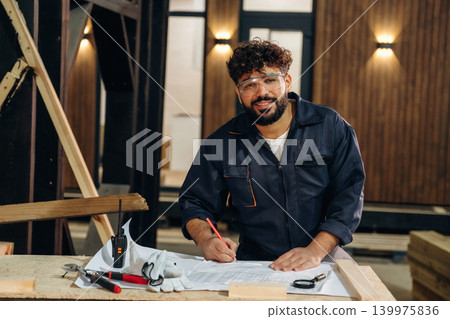 By the table with plan. Portrait of a male carpenter posing in a woodworking workshop 139975836
