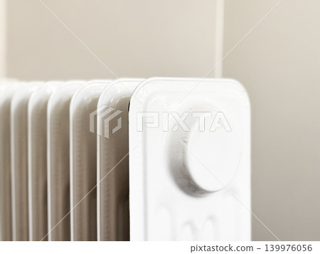 Closeup with of white heating radiator against light colored wall. Multiple sections with knob for adjusting heat level. Warming room during winter or colder days. House comfort in chilly weather Closeup with of white heating radiator against light colored wall. Multiple sections with knob for adjusting heat level. Warming room during winter or colder days. House comfort in chilly weather 139976056