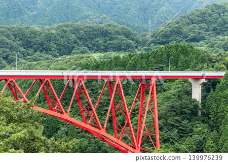 A landscape featuring a red inverted Langer Bridge spanning a deep green mountain valley. A landscape featuring a red inverted Langer Bridge spanning a deep green mountain valley. 139976239