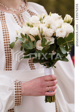 Bride in White Embroidered Dress Holding White Rose Bouquet with Green Leaves for Wedding Ceremony Bride in White Embroidered Dress Holding White Rose Bouquet with Green Leaves for Wedding Ceremony 139976856