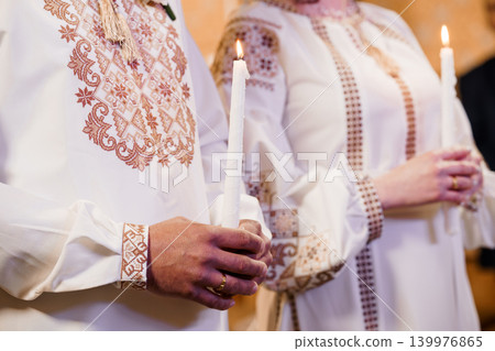 Bride and Groom in Traditional Embroidered White Outfits Holding Lit Wax Candles at Wedding Ceremony Bride and Groom in Traditional Embroidered White Outfits Holding Lit Wax Candles at Wedding Ceremony 139976865