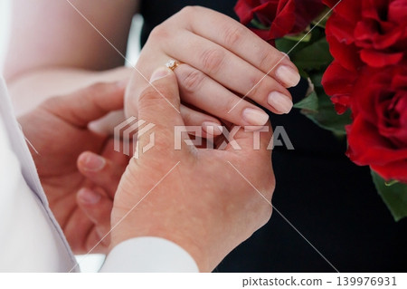 Close Up of Hands Placing Gold Diamond Engagement Ring on Woman Finger with Red Roses Close Up of Hands Placing Gold Diamond Engagement Ring on Woman Finger with Red Roses 139976931