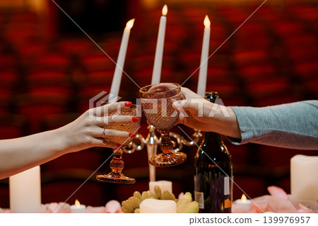 Two People Toasting with Pink Glassware and Wine in a Romantic Candlelit Theater Setting 139976957