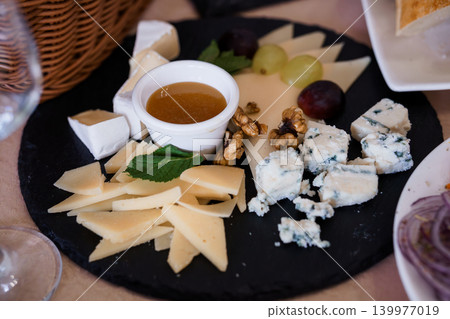 Assorted Cheese Platter with Grapes, Walnuts, and Honey Dip on Dark Slate Board 139977019