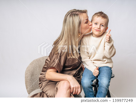 mom kisses boy 4-5 years old on the cheek, the boy smiles and shows a finger class, both European appearance blond boy with blue eyes, studio shooting, dressed in light clothes. High quality photo mom kisses boy 4-5 years old on the cheek, the boy smiles and shows a finger class, both European appearance blond boy with blue eyes, studio shooting, dressed in light clothes. High quality photo 139977249