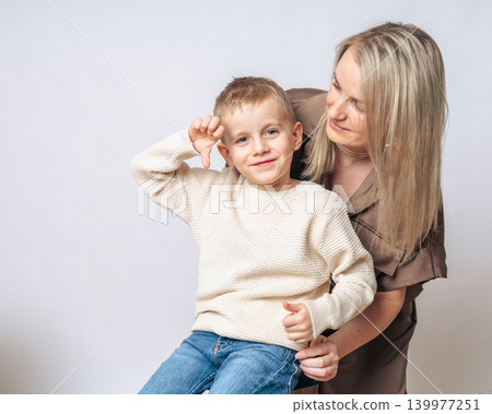 boy 4-5 years old boy sitting on mom's lap mom smiling and looking at the boy, the boy shows a finger down, both European appearance blond with blue eyes, studio shooting. High quality photo boy 4-5 years old boy sitting on mom's lap mom smiling and looking at the boy, the boy shows a finger down, both European appearance blond with blue eyes, studio shooting. High quality photo 139977251