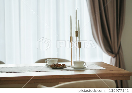 Gold candlesticks with white candles, two elegant white tea and coffee cups and walnuts on a table with a white tablecloth, curtains in the background Gold candlesticks with white candles, two elegant white tea and coffee cups and walnuts on a table with a white tablecloth, curtains in the background 139977341