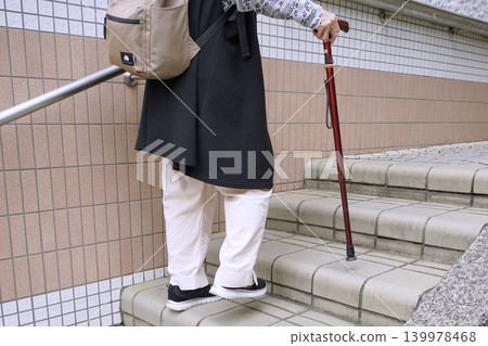 An elderly woman with a cane climbs the stairs in Yokohama, Japan, taking breaks along the city's urban landscape. An aging society. Even these gentle stairs become difficult for the elderly. 139978468
