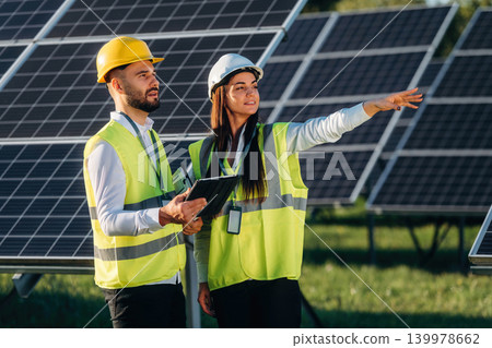 Communication between employees. Two solar engineers standing together at a photovoltaic field, in bright safety vests Communication between employees. Two solar engineers standing together at a photovoltaic field, in bright safety vests 139978662