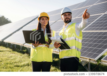 Laptop is in hands if woman. Two solar engineers standing together at a photovoltaic field, in bright safety vests Laptop is in hands if woman. Two solar engineers standing together at a photovoltaic field, in bright safety vests 139978674
