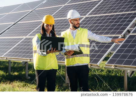 Laptop is in hands if woman. Two solar engineers standing together at a photovoltaic field, in bright safety vests Laptop is in hands if woman. Two solar engineers standing together at a photovoltaic field, in bright safety vests 139978676