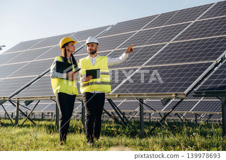 Explaining the plan and showing are to the woman. Two solar engineers standing together at a photovoltaic field, in bright safety vests Explaining the plan and showing are to the woman. Two solar engineers standing together at a photovoltaic field, in bright safety vests 139978693