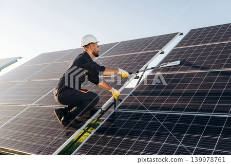 High up, cleaning the surface with a mop. Male solar engineer examining photovoltaic panels at a power station High up, cleaning the surface with a mop. Male solar engineer examining photovoltaic panels at a power station 139978761