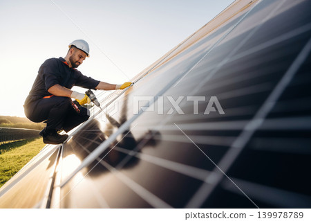 Service, sitting with drill and doing installation. Male solar engineer examining photovoltaic panels at a power station Service, sitting with drill and doing installation. Male solar engineer examining photovoltaic panels at a power station 139978789