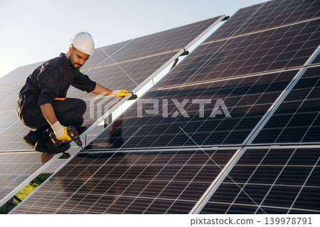 Service, sitting with drill and doing installation. Male solar engineer examining photovoltaic panels at a power station Service, sitting with drill and doing installation. Male solar engineer examining photovoltaic panels at a power station 139978791