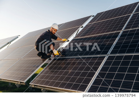 Service, sitting with drill and doing installation. Male solar engineer examining photovoltaic panels at a power station Service, sitting with drill and doing installation. Male solar engineer examining photovoltaic panels at a power station 139978794